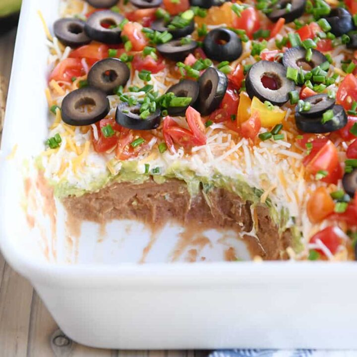 scoop taken out of 7-layer dip in white casserole dish