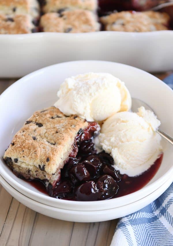 Scoop of cherry cobbler with chocolate chunk biscuit in white bowl with vanilla ice cream.