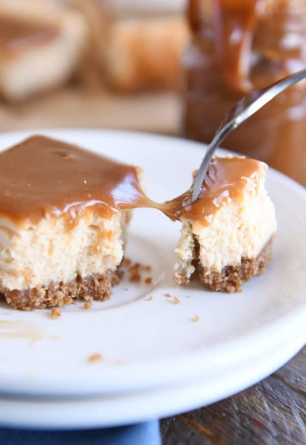 Cutting off bite of dulce de leche cheesecake square with fork on white plate.