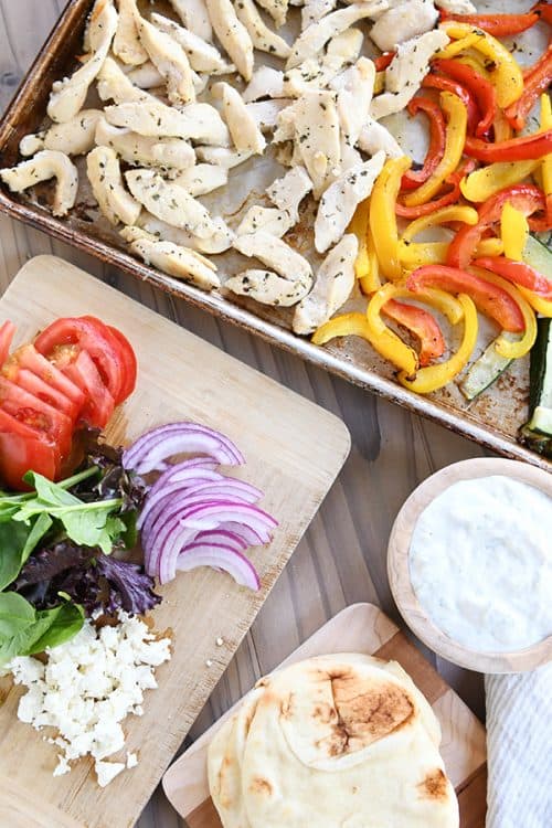 A sheet pan with cooked chicken and pepper slices next to a cutting board with sliced vegetables.