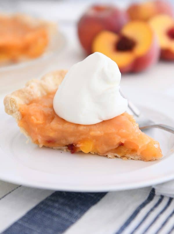 slice of fresh peach pie on white plate with spoonful of whipped cream on top
