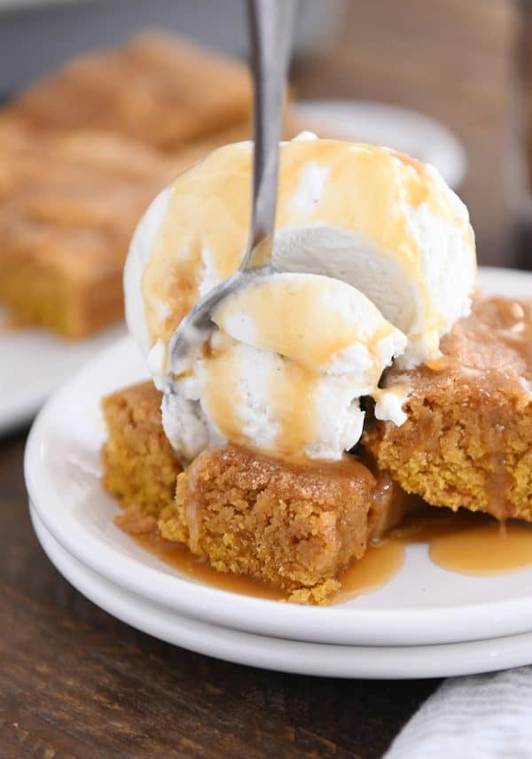 Fork taking bite out of easy pumpkin snickerdoodle bar with vanilla ice cream and butterscotch sauce on white plate.