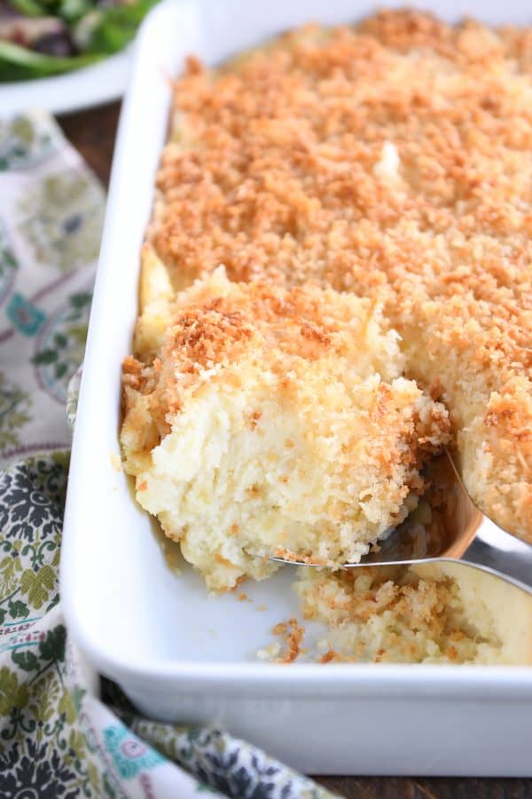 Metal spoon scooping out spoonful of baked mashed potatoes with parmesan topping in white dish.