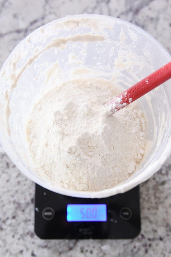 Adding flour to sourdough starter in bucket on black scale.