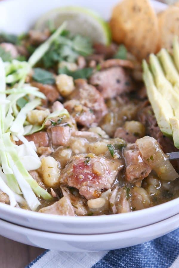 Seasoned pork, hominy, broth, cabbage, avocado in white bowl.