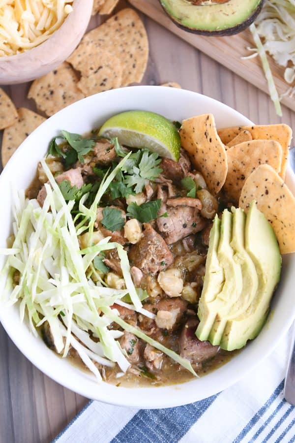 top down view of slow cooker posole in white bowl with shredded cabbage, sliced avocado, tortilla chips and lime wedge