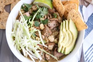 top down view of slow cooker posole in white bowl with shredded cabbage, sliced avocado, tortilla chips and lime wedge