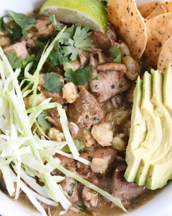 top down view of slow cooker posole in white bowl with shredded cabbage, sliced avocado, tortilla chips and lime wedge