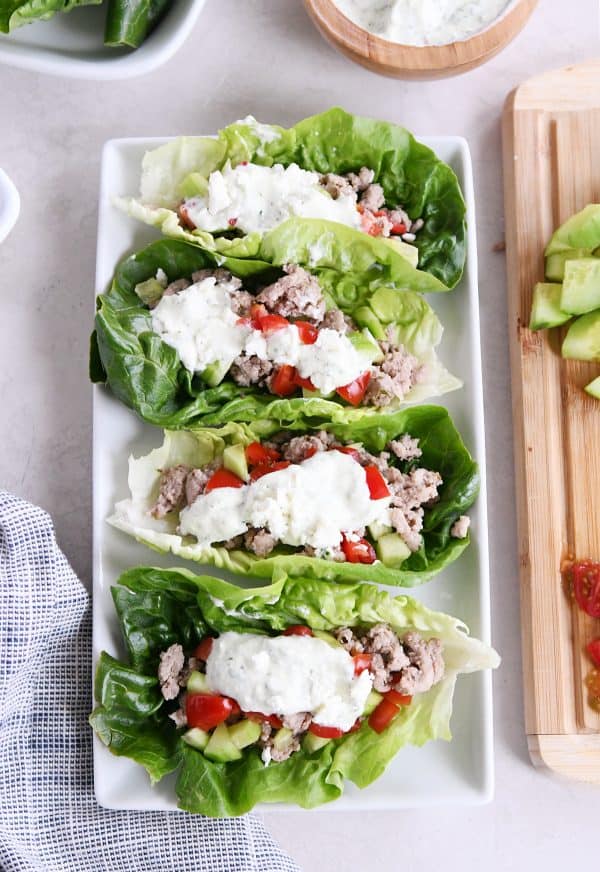 Four greek lettuce wraps on white tray with cucumbers, tomatoes, with feta mint dressing.