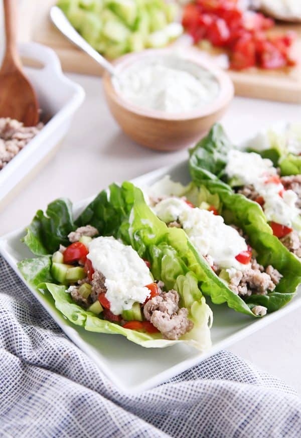 Three greek lettuce wraps on white tray with cucumbers, tomatoes, with feta mint dressing.