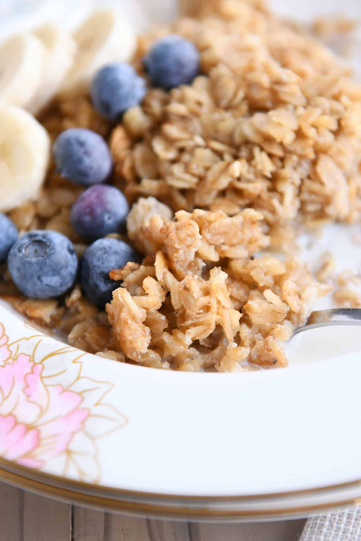 Spoon underneath spoonful of baked oatmeal, blueberries and sliced bananas in floral bowl.