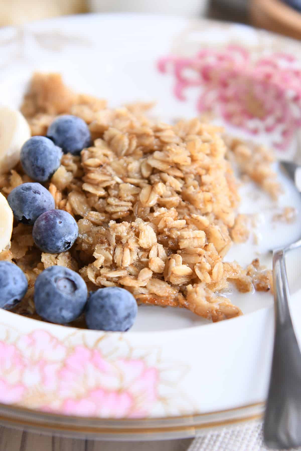 Baked oatmeal in floral bowl with spoon, milk, blueberries, and sliced bananas.