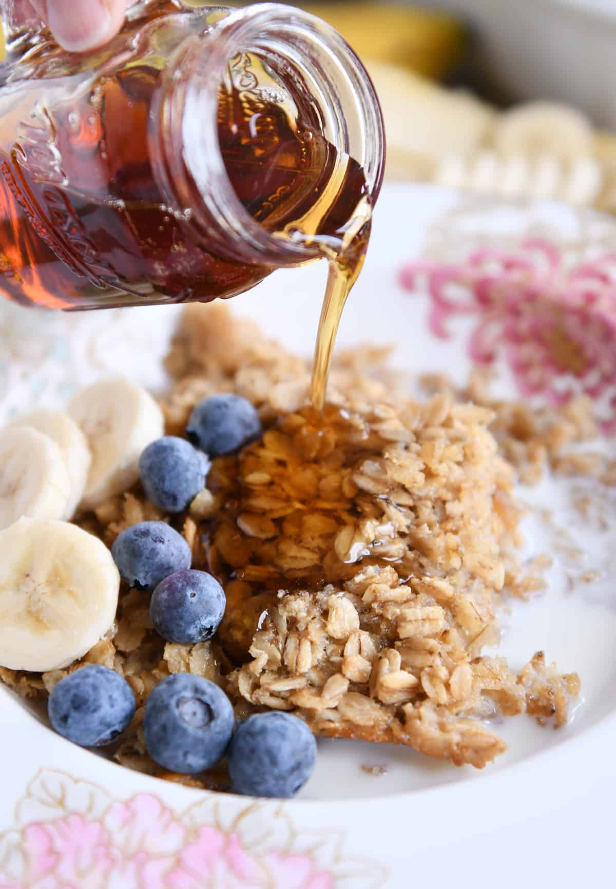pouring maple syrup over bowl of amish baked oatmeal with blueberries and sliced bananas