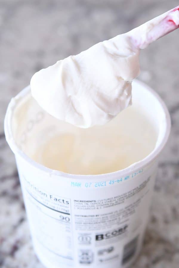 Spatula covered in plain yogurt hovering over yogurt tub.