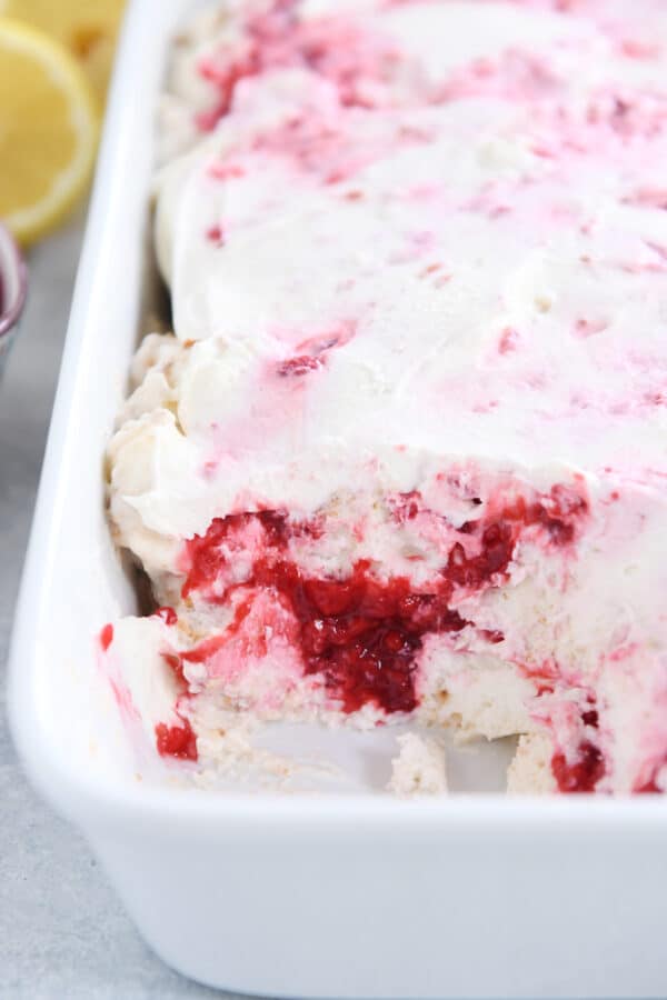 Layered angel food cake, raspberries and whipped cream in white baking dish with scoop taken out.