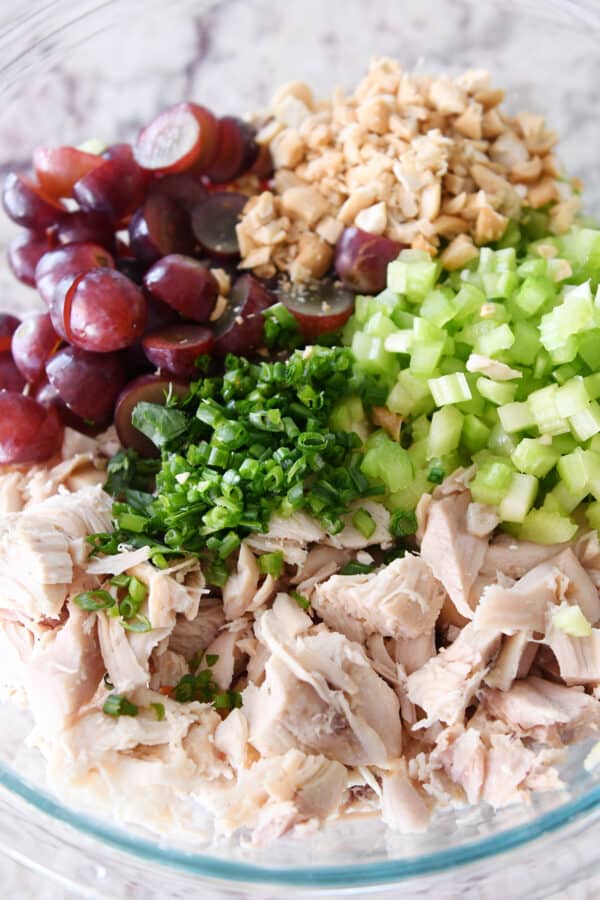 Shredded chicken, green onions, celery, grapes and nuts in glass bowl.