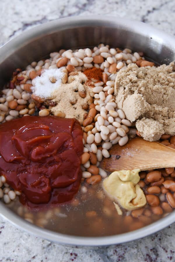 White beans, brown sugar, ketchup, broth, dijon mustard in shallow pan with wooden spoon.