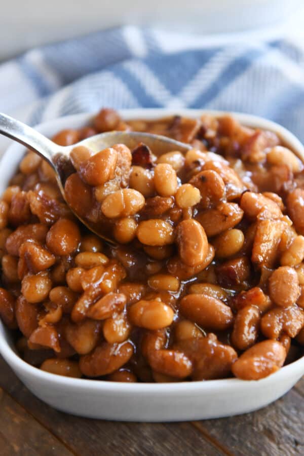 Spoon scooping out spoonful of saucy baked beans in white square dish.