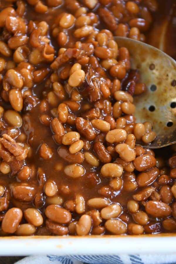 Slotted serving spoon scooping baked beans in white dish.