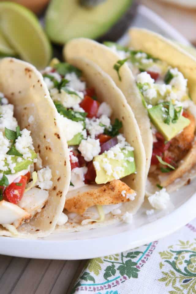 close up of three fish tacos on white platter with crumbled cheese, pico de gallo, and avocados