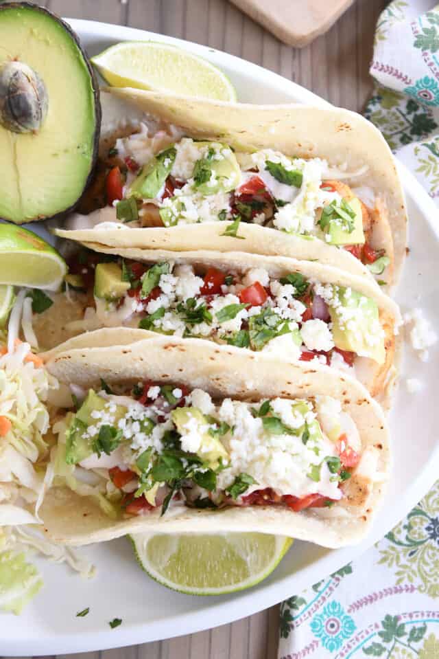 Three fish tacos on white platter with avocado and lime wedges.