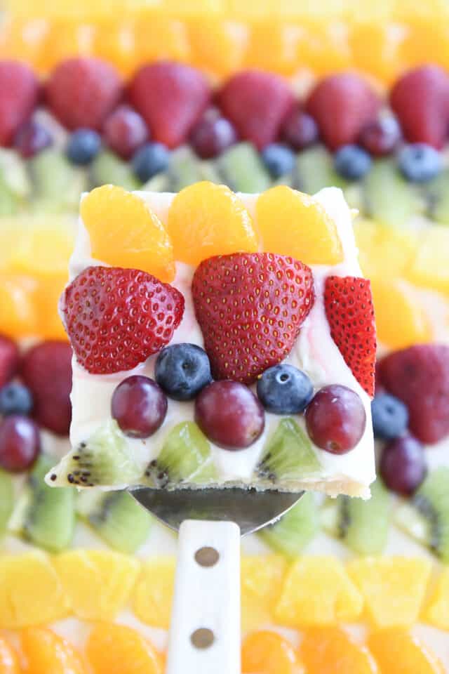 Square of fruit pizza with mandarin oranges, strawberries, blueberries, red grapes and kiwi above sheet pan fruit pizza.