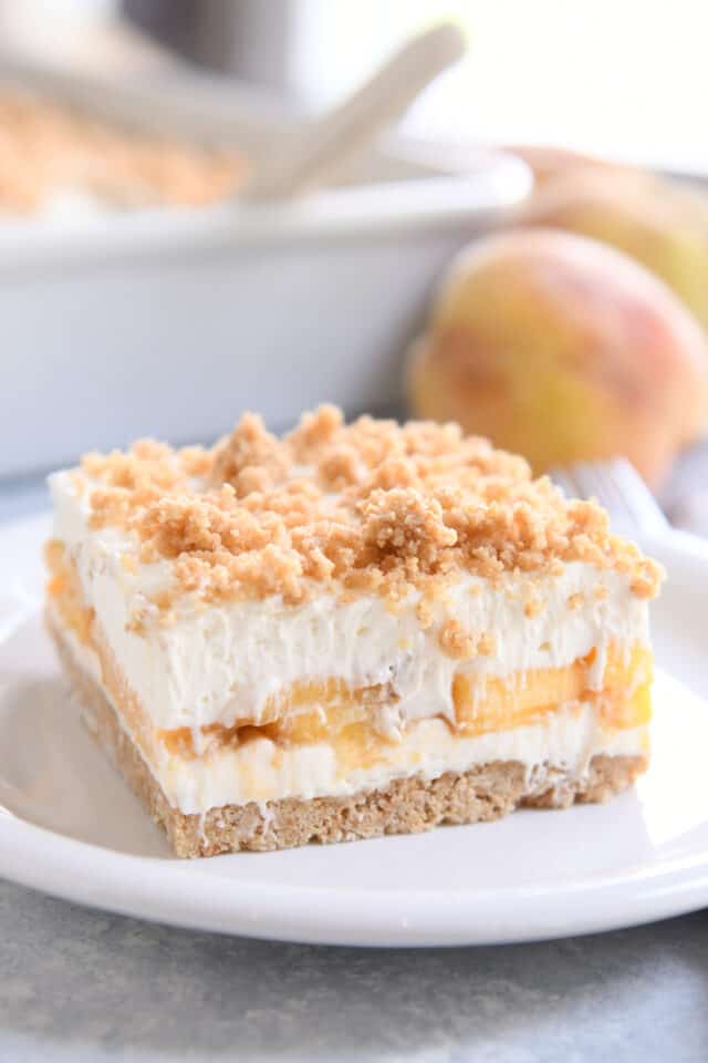 Square of peaches and cream dessert on white plate with peach in background.