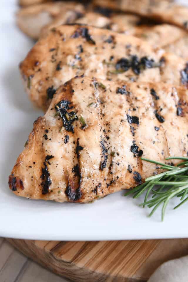 Grilled chicken breasts on white platter with fresh rosemary sprig.