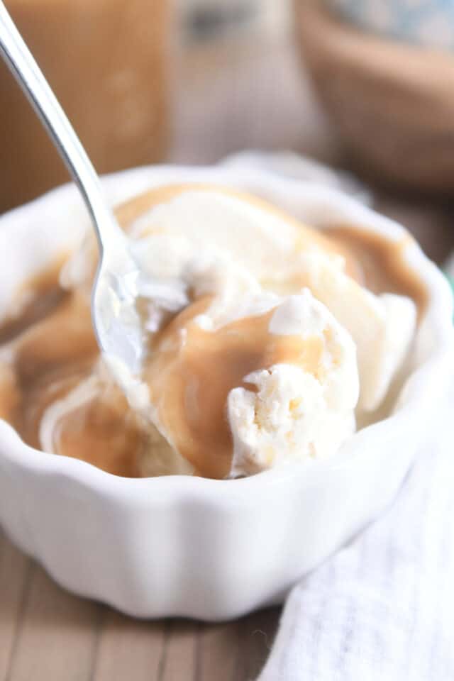 Spoon scooping vanilla ice cream and butterscotch sauce out of white bowl.