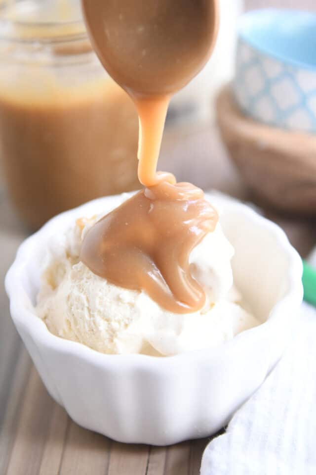 Wooden spoon drizzling butterscotch sauce over vanilla ice cream in white bowl.