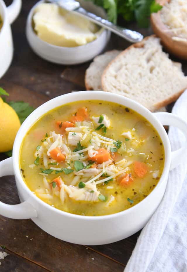 Lemon chicken orzo soup in white bowl with handles with slices of bread and butter.