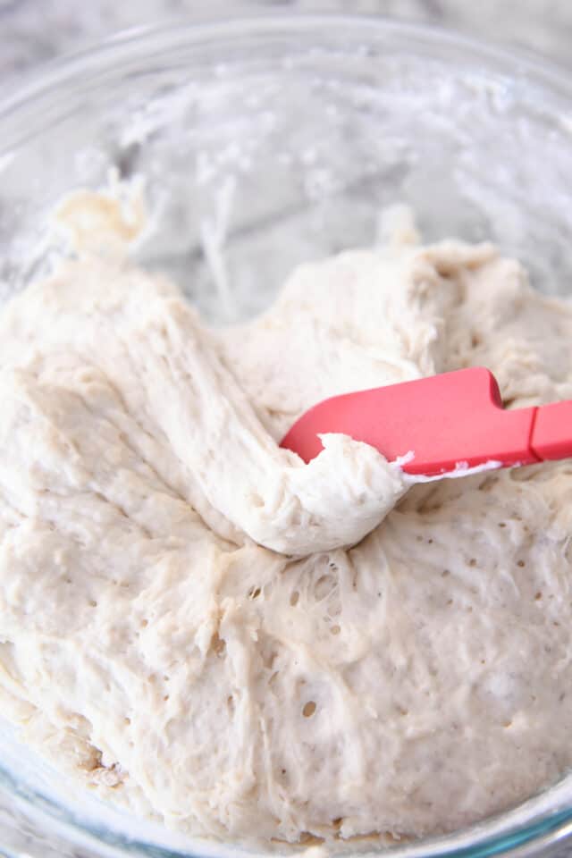 Peasant bread dough being scraped away from sides of glass bowl with rubber spatula.