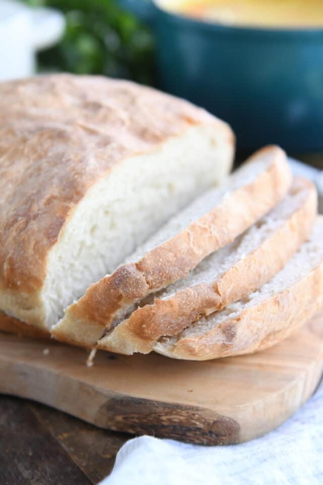 Loaf of no-knead peasant bread on wood cutting board with three slices cut.