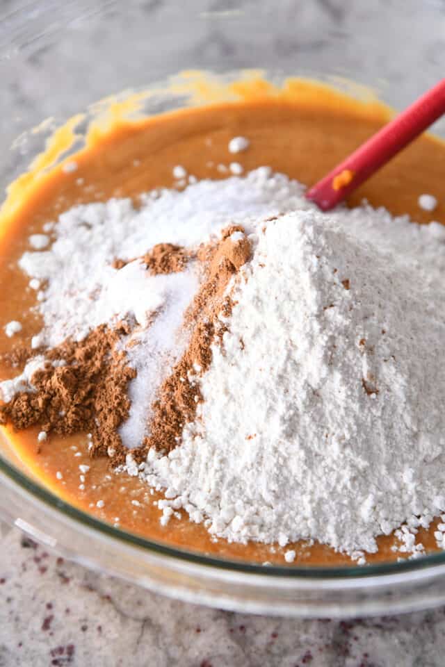 Adding flour, cinnamon and salt to wet ingredients for pumpkin muffin batter in glass bowl.