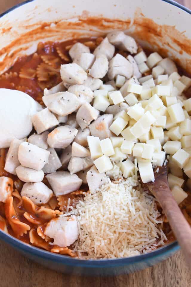 Mozzarella cubes, cooked chicken, parmesan cheese and ricotta cheese with bowtie pasta in cast iron pot.