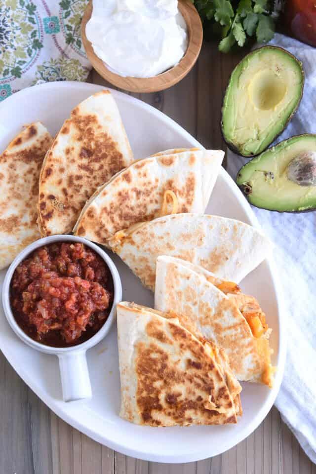 White platter of six chicken quesadilla triangles with a bowl of salsa, a cut avocado and a small wooden bowl of sour cream.