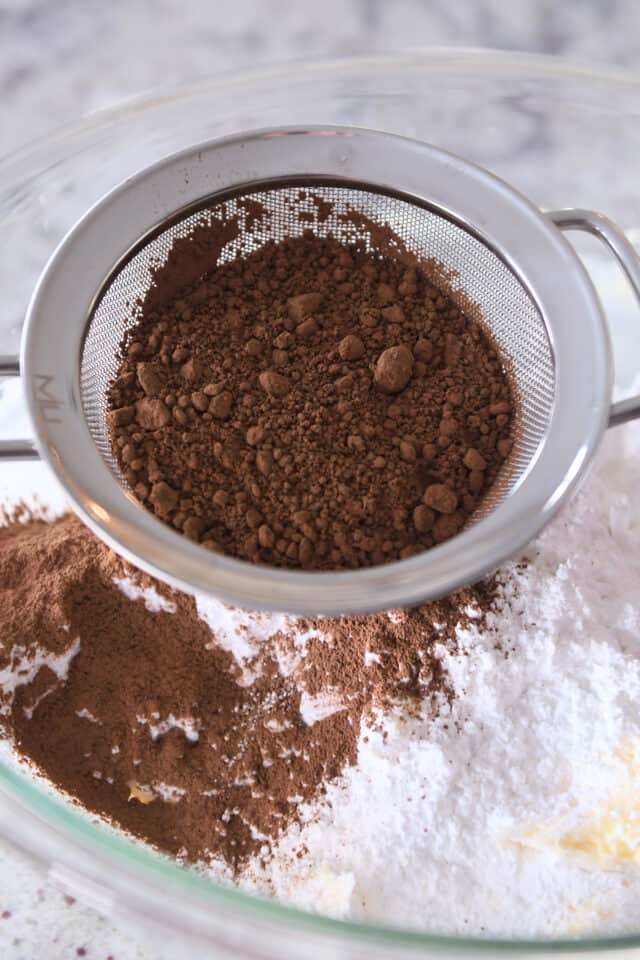 Sifting cocoa powder into glass bowl with powdered sugar and butter.