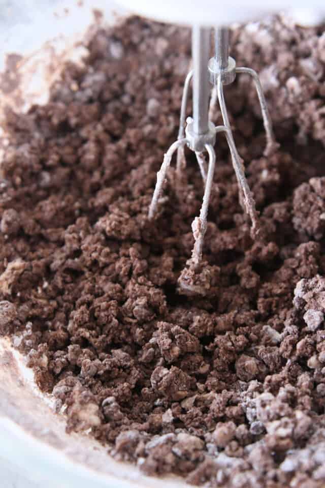 Mixing dry ingredients into chocolate frosting with handheld electric mixer.