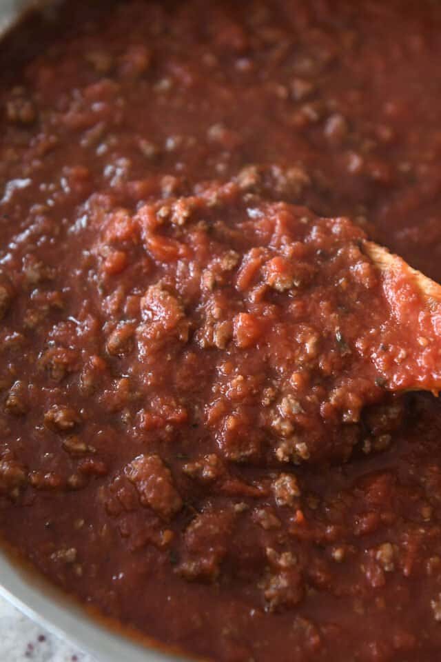 Wooden spoon scooping red sauce with ground beef in stainless pan.