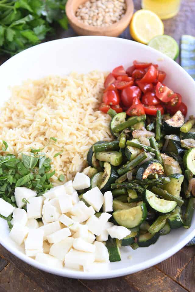 White bowl with cooked orzo, fresh basil, cherry tomatoes, roasted asparagus, and fresh mozzarella cheese.