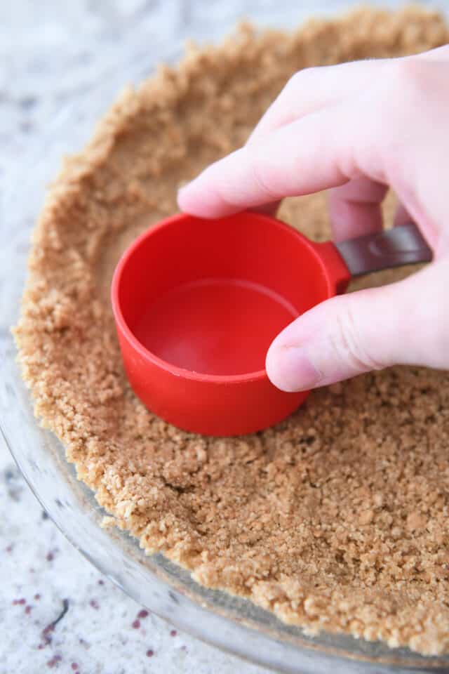 Pressing graham cracker crust into glass pie plate with measuring cup.