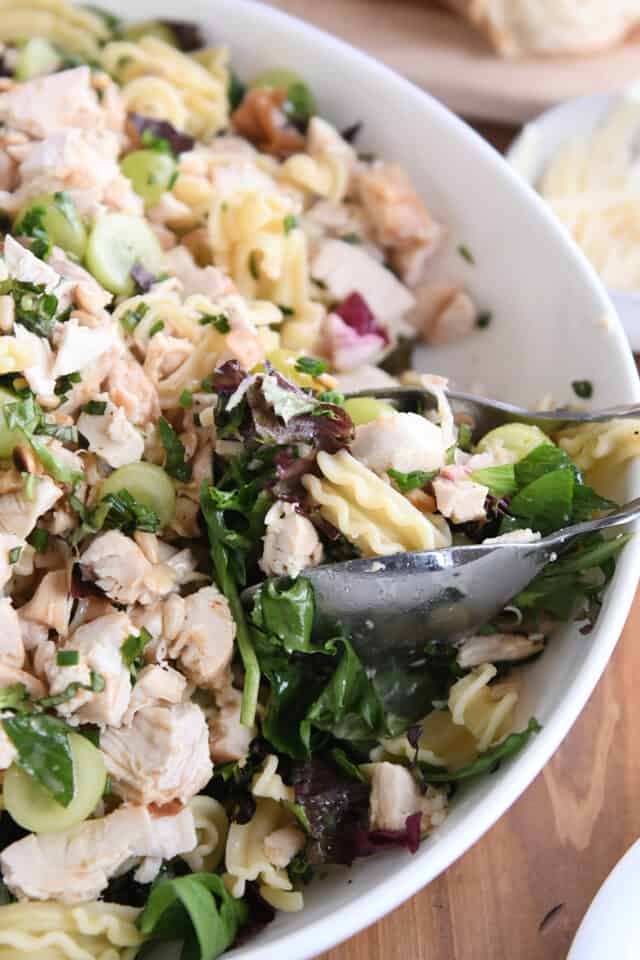 Scooping vineyard chicken and pasta salad with salad tongs out of white bowl.