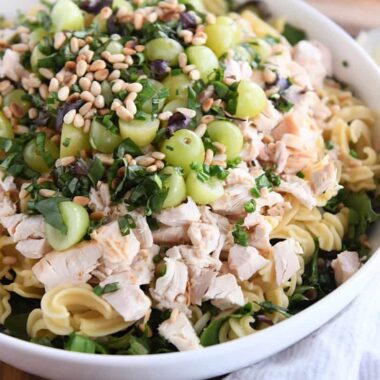 large white bowl with greens, pasta, chicken, grapes, fresh basil, and pine nuts