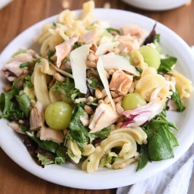 small white plate with greens, pasta, green grapes, chicken and pine nuts