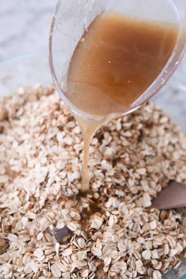 Pouring granola syrup over oats in glass bowl.