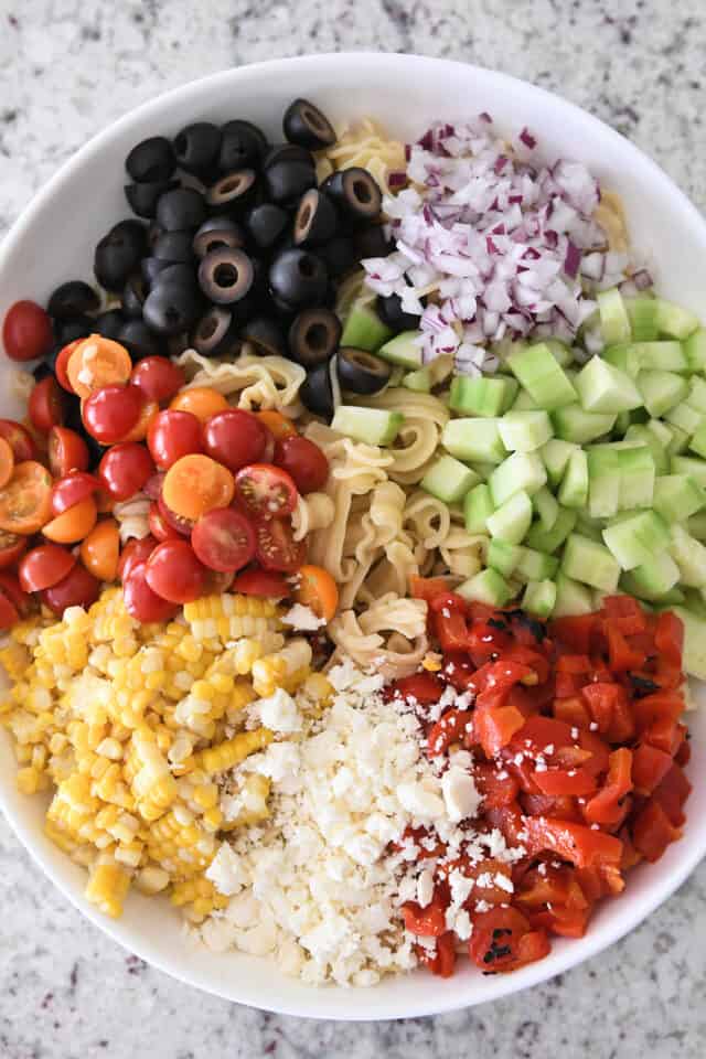 Top down view of feta cheese, corn, tomatoes, olives, red onions, cucumbers, red peppers and pasta.