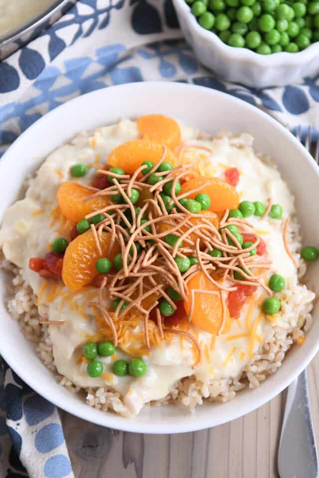 White bowl with brown rice, hawaiian haystacks gravy, mandarin oranges, chow mein noodles and peas.