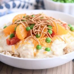 White bowl with brown rice, hawaiian haystacks gravy, mandarin oranges, chow mein noodles and peas.