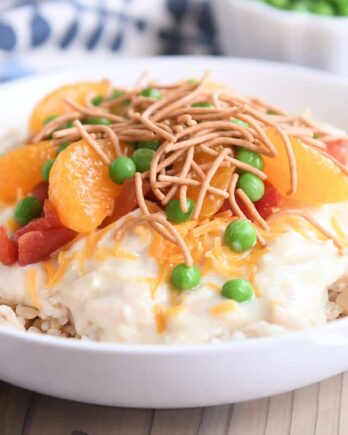 White bowl with brown rice, hawaiian haystacks gravy, mandarin oranges, chow mein noodles and peas.