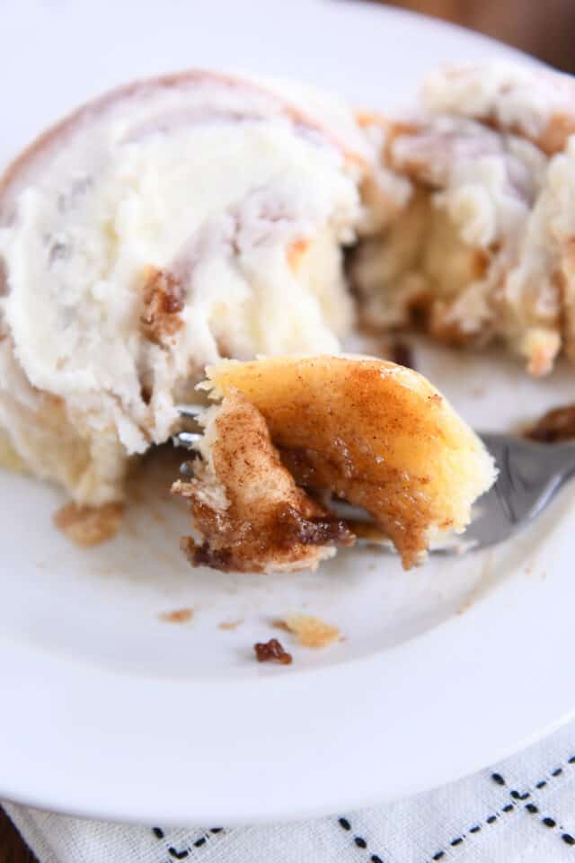 Bite of gooey cinnamon roll on fork on white plate.